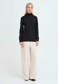 Schwarzer Rollkragenpullover mit langen Ärmeln, weicher Textur, figurbetonter Silhouette. Getragen mit lockeren cremefarbenen weiten Hosen und schwarzem Schuhwerk.