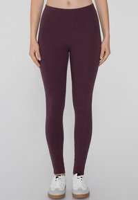 Burgundské legíny vyrobené z elastického materiálu s hladkým povrchem, s vysokým pasem a úzkým střihem, doplněné bílými sportovními botami.
