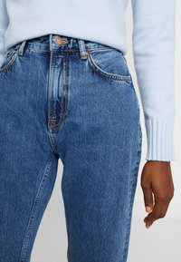 Högmidjade denimjeans i en mellanblå tvätt med oranga sömmar, femfickorsdesign och kopparknappslås. Strukturerad tyg.