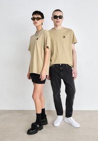 Kaksi mallia seisoo vierekkäin. Naisella on päällään beige ylisuurennettu t-paita ja musta minihame, lisäksi hänellä on platform-kengät. Miehellä on päällään beige t-paita ja mustat farkut, joissa on valkoiset lenkkarit.