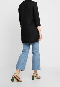 Musta tunika paita, jossa on kolme neljännestuntia hihat, yhdistettynä leveälahkeisiin sinisiin farkkuihin. Vihreät remmilla varustetut, paksupohjaiset sandaalit, joissa on teksturoitu pinta.