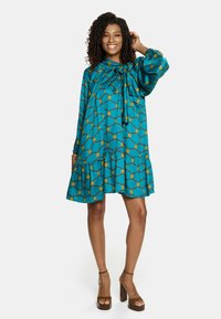 Turkusowa sukienka z dekoltem w kształcie kokardy, długimi rękawami i rozkloszowanym dołem. Ozdobiona geometrycznym wzorem w kolorze żółtym i niebieskim. Gładka tekstura materiału.