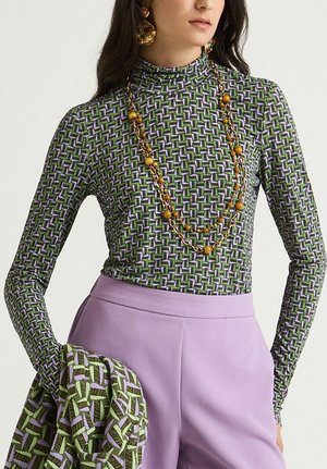 Top verde a maniche lunghe e aderente con motivo geometrico viola e bianco. Abbinato a pantaloni lavanda e tessuto stampato drappeggiato sul braccio.