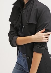 Musta trench-coat, jossa on taitettu kaulus, napitettu etuosa ja käännetyt hihat. Ominaisuuksiin kuuluu sileä kangas ja istuva leikkaus.