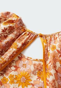 Blouse florale avec des motifs orange et jaune, tissu doux, encolure froncée et détail de dos en forme de clé maintenu par un noeud.
