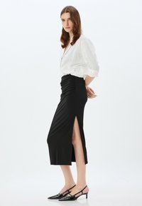 Jupe midi noire froncée avec fente sur le côté, associée à une chemise blanche à boutons. Des escarpins noirs à talons ouverts et à bouts pointus complètent le look.