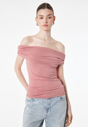 Jeune femme portant un haut rose épaules dénudées et un jean bleu clair, debout avec une main en poche devant un fond blanc.