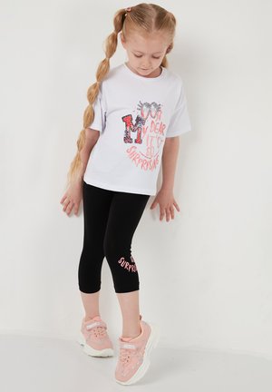 Jeune fille aux longs cheveux tressés portant un t-shirt blanc à motifs, des leggings noirs avec des inscriptions, et des baskets roses, debout contre un mur blanc.