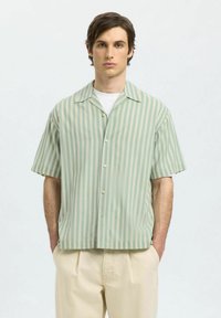 Camisa de botones de manga corta en verde claro con rayas verticales color crema. Tejido suave, corte relajado y cuello puntiagudo. Combinada con pantalones beige.