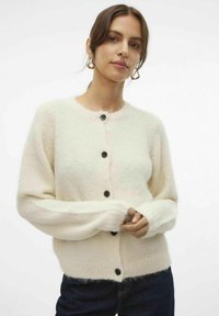 Witte, fluffy cardigan met ronde halslijn, zwarte knopen aan de voorkant en lange mouwen. Zachte textuur, relaxed fit, gecombineerd met donkere jeans.
