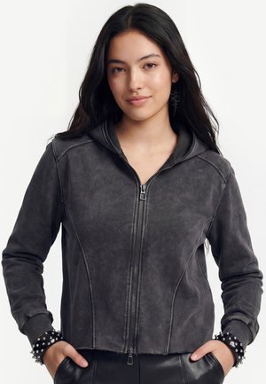 Jeune femme aux longs cheveux foncés portant un sweat à capuche gris foncé zippé avec des bracelets noirs cloutés, les mains dans les poches et une expression neutre.