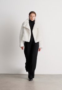 Λευκό ψευδές γούνινο μπουφάν cropped με φαρδύ γιακά, φορεμένο πάνω από μαύρο τζέρσεϊ και μαύρα παντελόνια. Λείος υφασμάτινος θρόισμα με χαλαρή εφαρμογή.