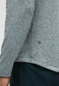 Šedé melírované tričko s dlhým rukávom s jemnou textúrou, zaobleným spodným okrajom a malým logom na boku, skombinované s tmavými nohavicami.