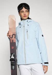 Gaiši zils slēpošanas jaku ar kapuci, ar rāvējslēdzēja priekšpusi un melniem akcentiem. Modelis tur snovbordu ar teksturētu virsmu un regulējamiem stiprinājumiem.