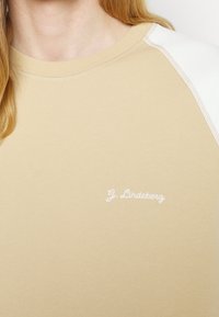 Sudadera de cuello redondo beige con acentos en blanco en los hombros y logotipo bordado en blanco. Textura suave, diseño casual y detalles cosidos.