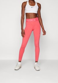 Žena v bílém sportovním topu, korálově růžových legínách Nike a bílých teniskách stojí na jednolitém bílém pozadí.
