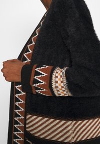 Osoba oblečená v černém chlupatém kardiganu s geometrickým vzorem hnědé, bílé a černé pletené výšivky na rukávech a okrajích.