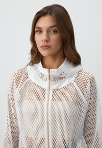 Giacca bianca in rete con zip e cappuccio, caratterizzata da un colletto liscio e hardware visibile, progettata con un motivo aperto e sciolto per una buona traspirazione.