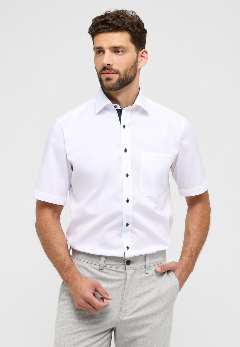 Camicia bianca a maniche corte con bottone frontale e bottoni blu scuri, dotata di taschino sul petto. Il materiale appare liscio e leggero.