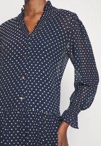 Mörkblå klänning med vita prickar, knappad framtill, volangkrage och långa plisserade ärmar med elastiska muddar på personens överkropp.