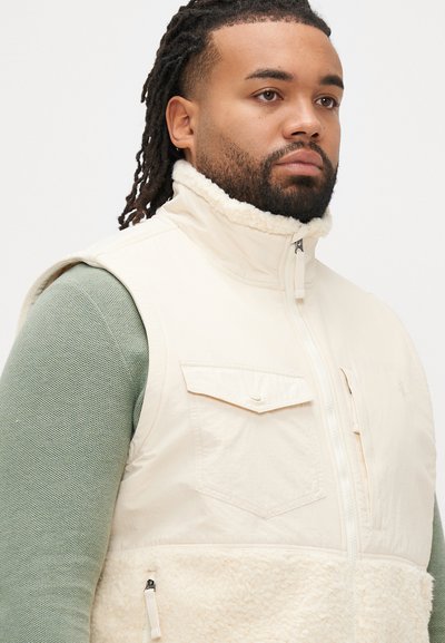 Colete bege com textura fofa de lã, fecho de correr e dois bolsos frontais. Usado sobre uma camisa de manga comprida verde clara, com gola alta.