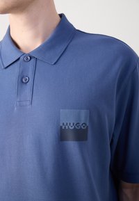 Blå polo skjorta med en texturerad tyg, en tvåknapps knäppning och en framträdande fyrkantig logotyp på vänster bröst med texten "HUGO".