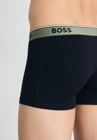 Schwarze Boxer Shorts mit einem grünen elastischen Bund, der das „BOSS“ Logo trägt. Glatte Textur, enganliegendes Design und mittellange Oberschenkel.