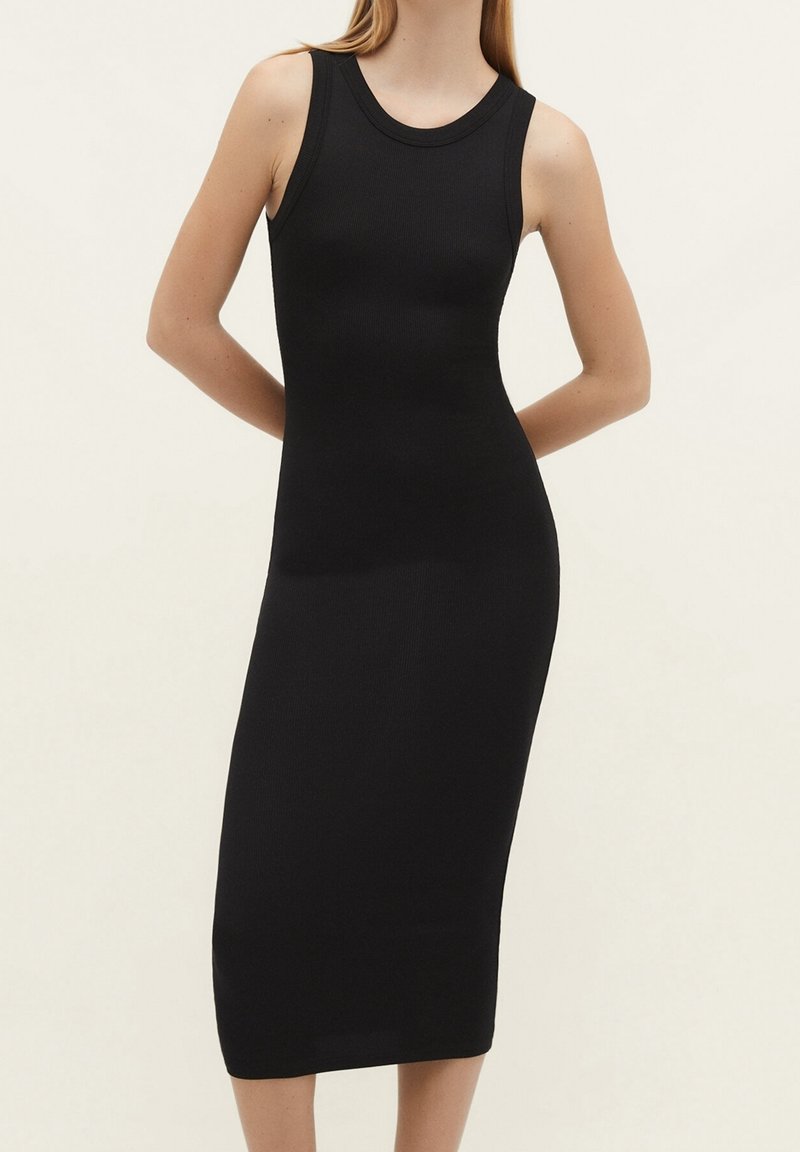 Femme portant une robe midi noire ajustée sans manches avec un col rond, debout, les mains derrière le dos, devant un fond clair uni.
