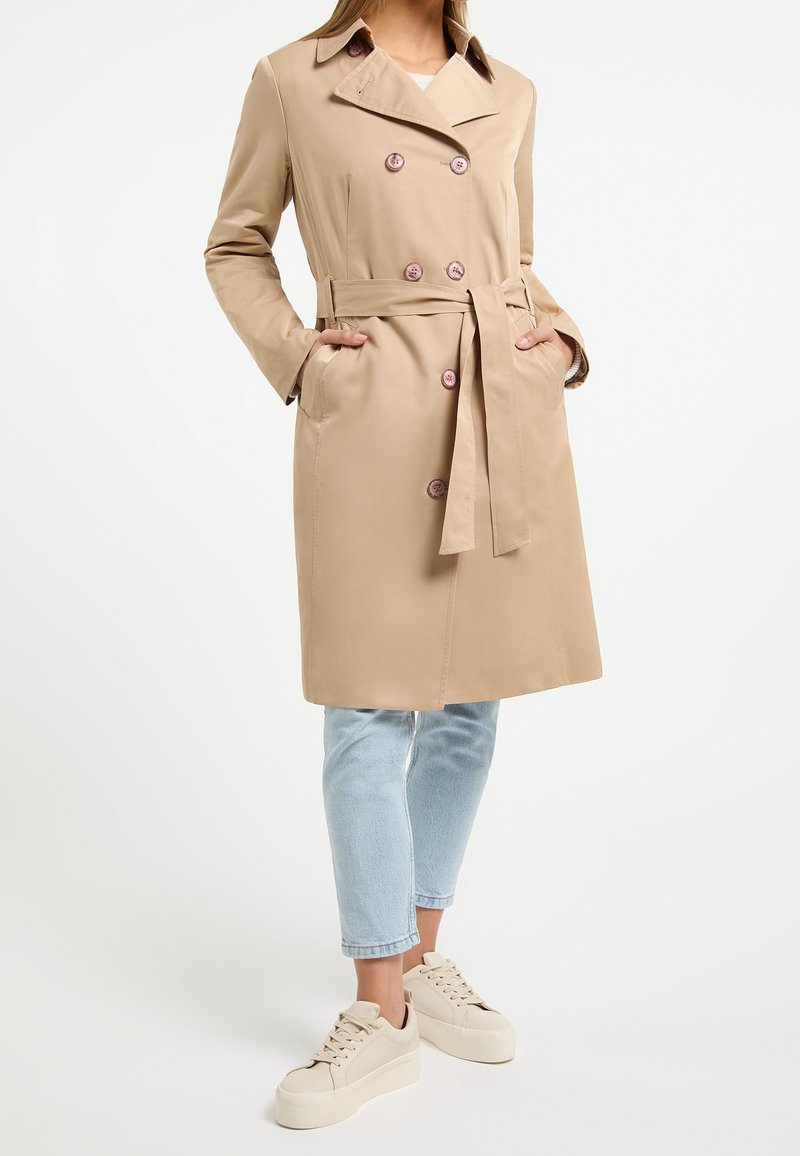 Femme portant un trench-coat beige longueur genoux avec une ceinture nouée à la taille, un jean court bleu clair et des baskets compensées beiges.