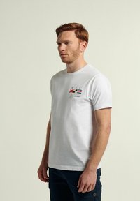 Camiseta de algodón blanca con mangas cortas, que presenta logotipos bordados coloridos en el pecho, incluyendo texto y símbolos náuticos.