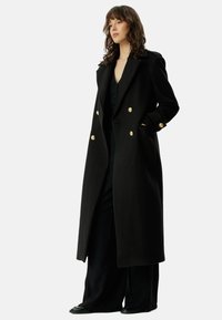 Black wool overcoat with a double-breasted design, gold button accents, wide lapels, and side pockets, worn over a black ensemble.