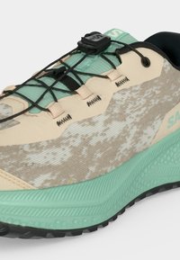Gros plan d'une chaussure de sport beige et verte avec un système de laçage rapide noir et une semelle texturée sur un fond clair.