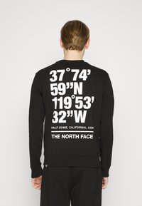 Svart sweatshirt med stor vit text på baksidan som visar koordinater och platsinformation. En enkel design med rund halsringning och ribbade manschetter.