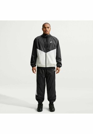 Hombre de pie vestido con un chándal Nike blanco y negro con chaqueta con capucha y pantalones a juego, frente a un fondo claro y liso.