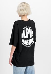 Černé oversized tričko s bílým grafickým textem "ALPHA INDUSTRIES" a "SINCE 1959" na zadní straně. Bavlněný materiál, krátké rukávy, volný střih.