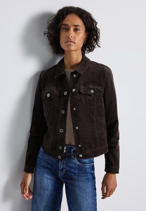 Woman with curly hair wearing a dark brown buttoned jacket, gray shirt, and blue jeans, standing against a plain light background.