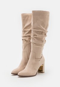 Stivali beige al ginocchio in morbido suede, caratterizzati da un design slouchy, punta a punta e un tacco a blocco con dettagli dorati.