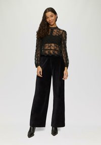 Schwarzes Spitzen-Cropped-Longsleeve-Top mit gewelltem Saum, kombiniert mit hochtaillierten, schwarzen Samtweitbeinhosen und spitzen, schwarzen Ankle Boots.