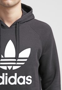 Svart bomullshoodie med en stor vit Adidas-logga, med dragkedja i huvan och raglanärmar. Mjuk textur, minimalistisk design.