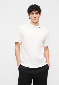 Νεαρός άνδρας με σγουρά σκούρα μαλλιά, φοράει λευκό κοντομάνικο μπλουζάκι και μαύρο παντελόνι, με τα χέρια στις τσέπες, κοιτάζοντας μπροστά σε λευκό φόντο.