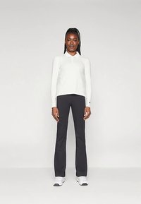 Mulher de pé, de frente, vestindo uma camisa branca de manga longa com colarinho, calças pretas e ténis brancos, em um fundo simples.