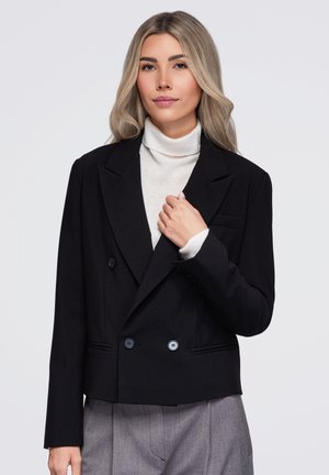 Femme aux longs cheveux blonds porte un blazer noir, un col roulé blanc et un pantalon gris, tenant le revers du blazer d'une main sur fond blanc.