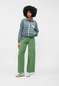 Μπλε φερμουάρ μπουφάν με μοτίβο, λευκή τσέπη; πράσινα καπιτονέ παντελόνια σε φαρδιά γραμμή; ανοιχτόχρωπα παπούτσια με λουριά και κάλτσες.