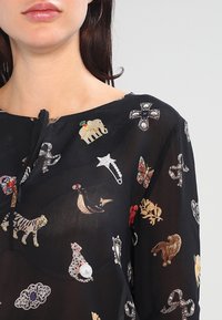 Schwarze transparente Bluse mit verschiedenen dekorativen Verzierungen, einschließlich Tier- und Schmuckmotiven, runder Halslinie und Kordelzugdetail.