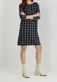 Abito a quadri blu navy con maniche a tre quarti, dotato di scollatura quadrata e taglio dritto. Indossato con stivaletti alla caviglia color crema.