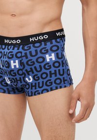 Blauwe boxershorts met zwart-wit "HUGO" logo patroon, voorzien van een zwarte tailleband met witte tekst. Zacht, rekbaar materiaal.
