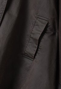 Tissu marron texturé avec un léger éclat, présentant une poche latérale avec un rabat replié. Des variations subtiles de couleur ajoutent de la profondeur.