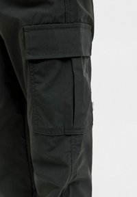 Un pantalon cargo noir montrant une grande poche latérale avec un rabat et des coutures visibles sur un fond clair.