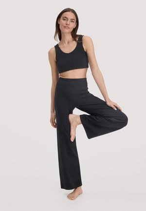 Vrouw in zwarte mouwloze crop top en wijde broek die op één been balanceert tegen een effen lichte achtergrond.