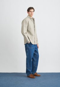 Camisa de botones color beige claro con mangas largas, combinada con unos vaqueros de denim azul de corte holgado y zapatos marrones de cordones. Diseño simple y ajuste relajado.